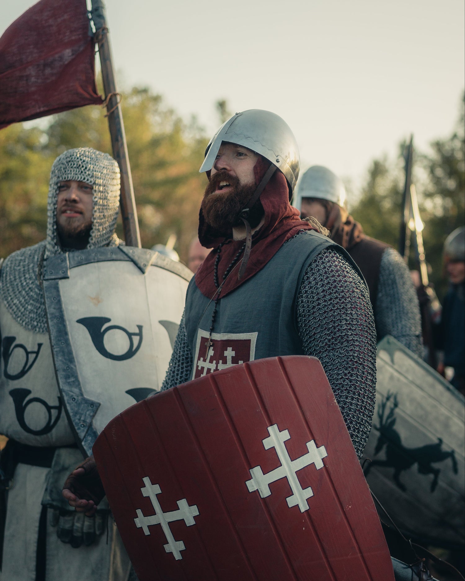 knight in medieval armor holding a red shield with white cross design, surrounded by other similarly dressed individuals.